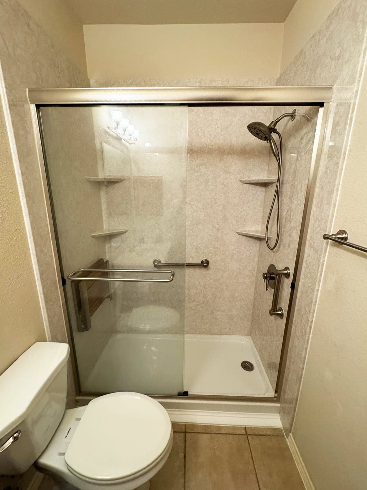 A remodeled walk-in shower with stone-look wall panels, corner shelves, a grab bar, a handheld shower on a slide bar, and sliding glass doors